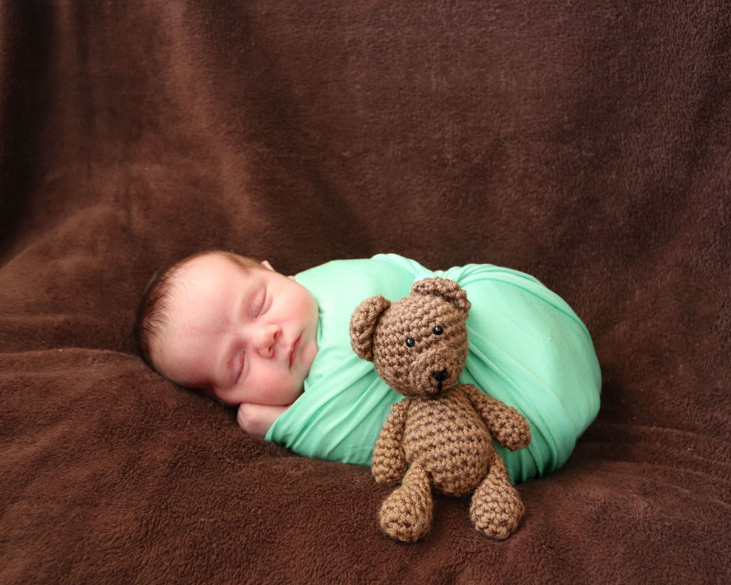 Peaceful sleeping baby wrapped in mint green swaddle with brown teddy bear on brown backdrop.