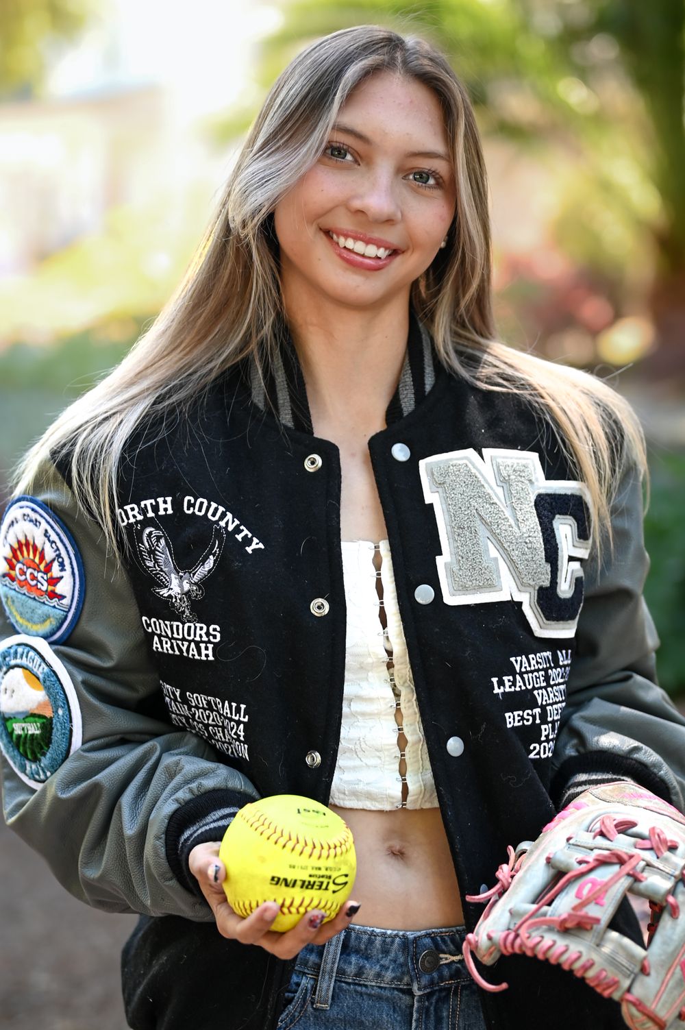 Softball player in varsity jacket holds yellow ball and glove outdoors.