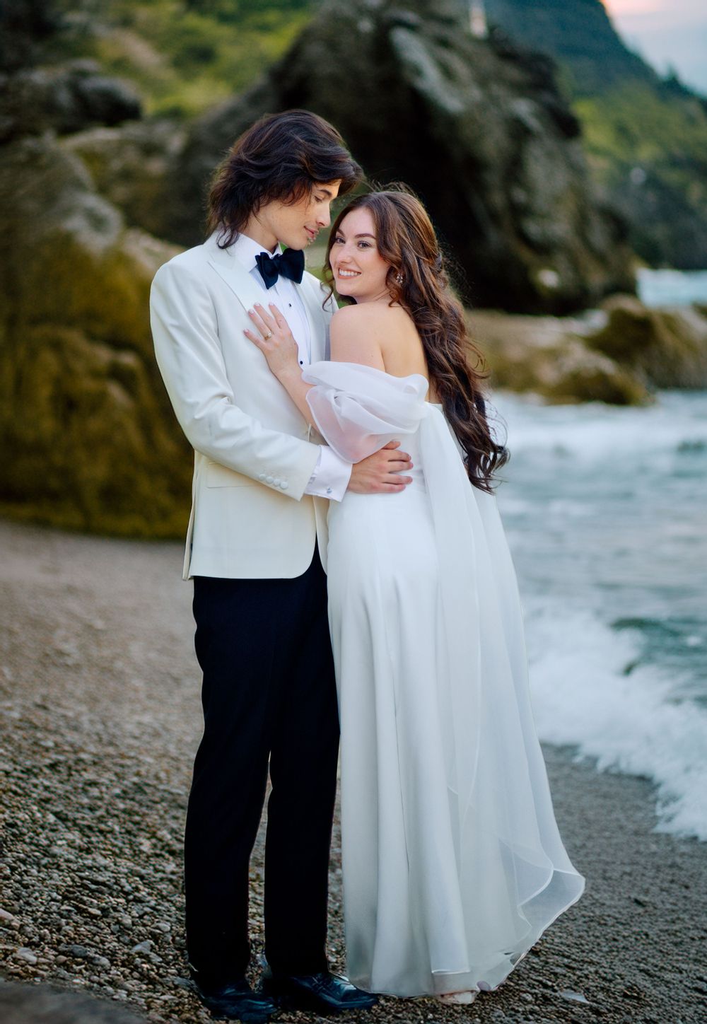 A romantic wedding couple embraces on a rocky beach with mountains and waves in the background at sunset.