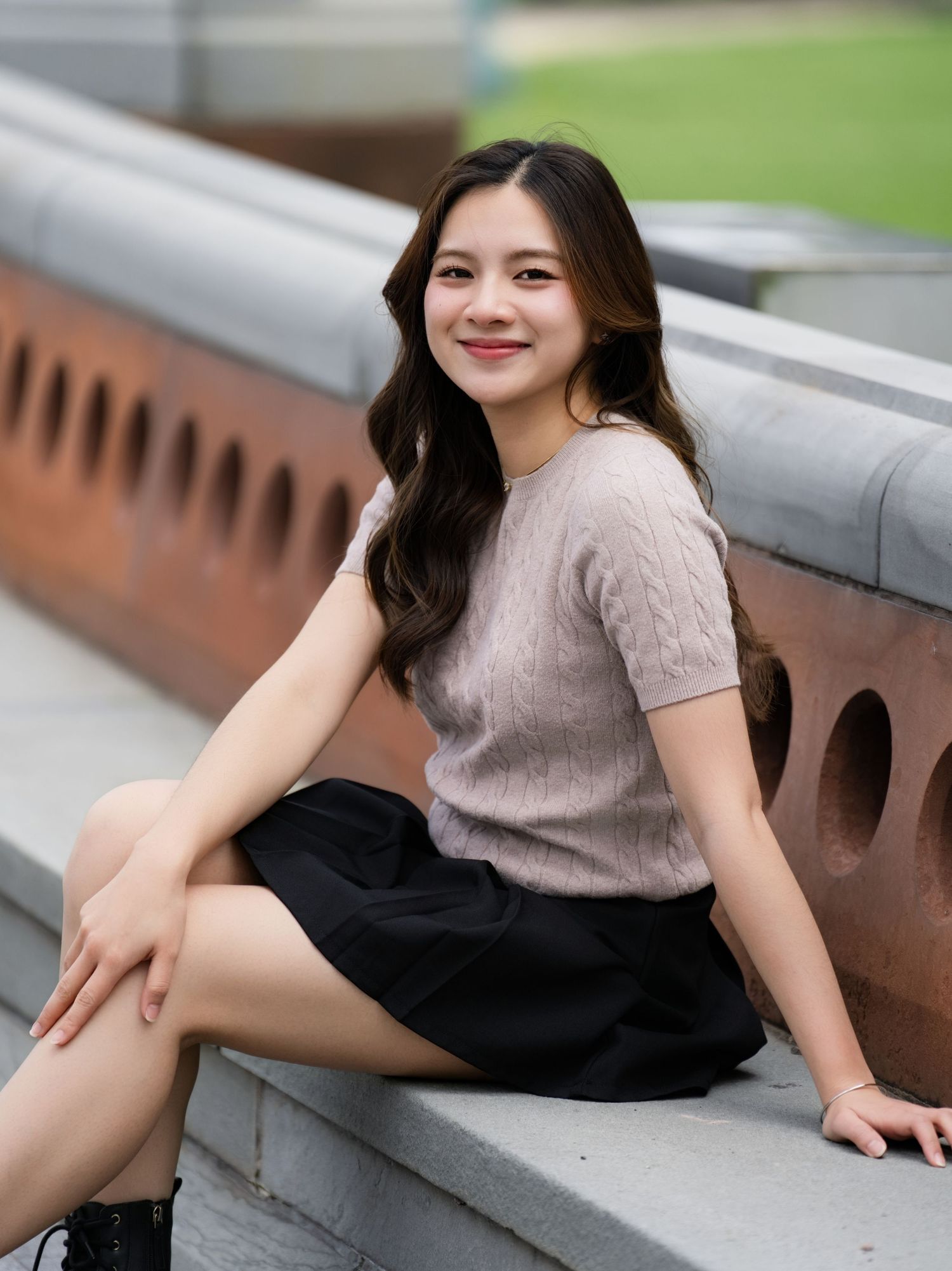 A person in a pink short-sleeved top and black skirt poses casually on concrete steps outdoors.