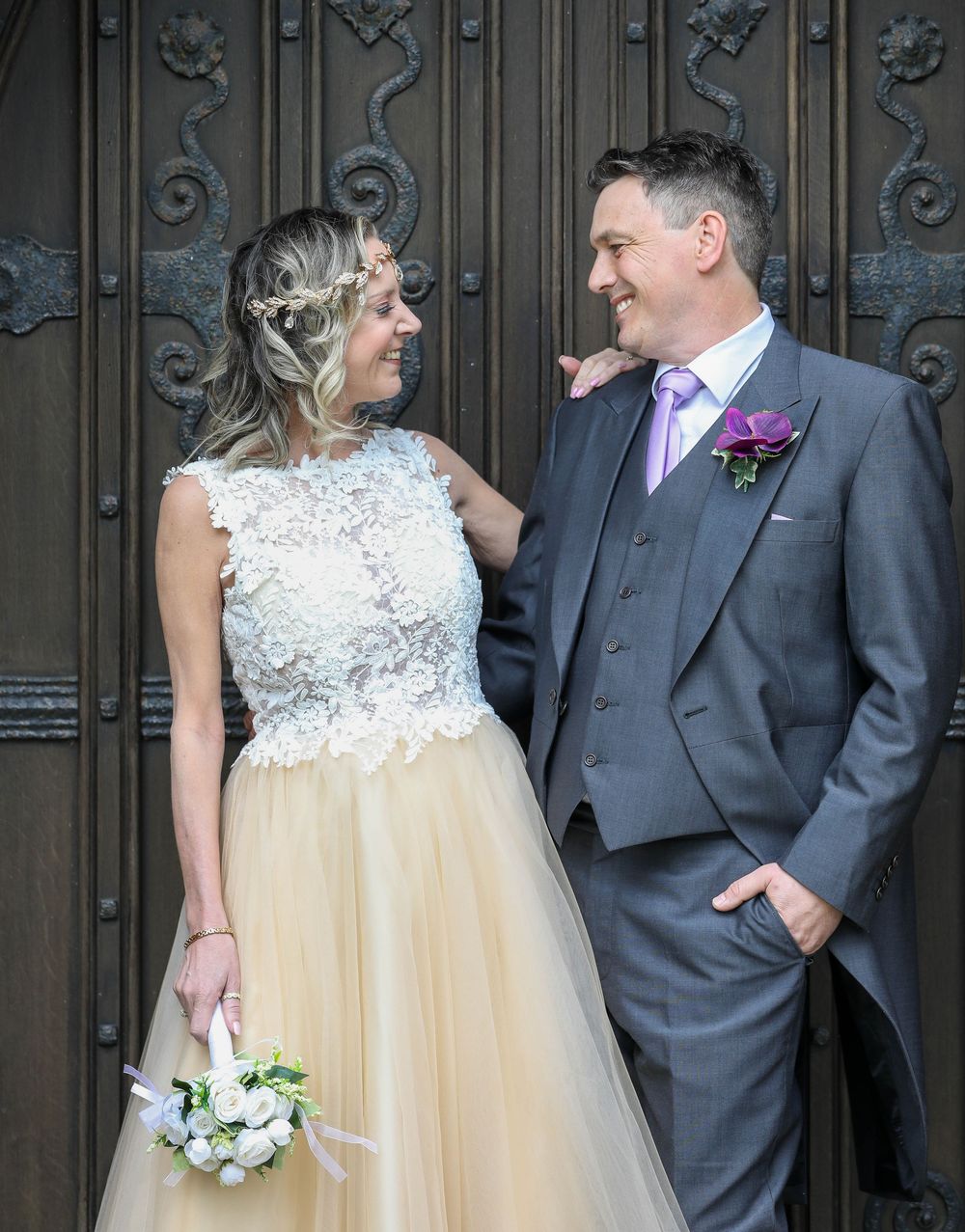 Wedding couple poses against ornate metal door showing elegant dress and suit details.
