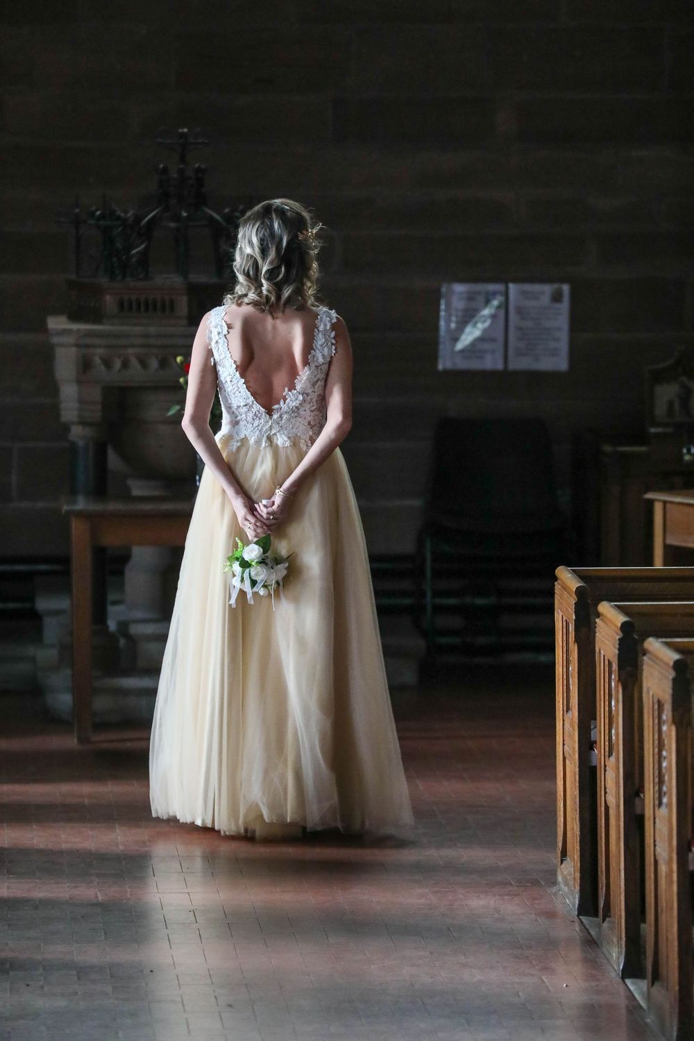 Bride in champagne tulle gown stands in church aisle holding white bouquet.