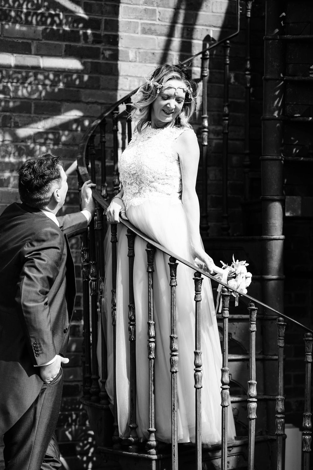 Black and white photo of bride in lace dress posing on vintage staircase with groom below.