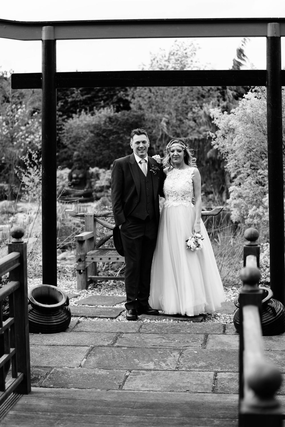 Black and white portrait of newlyweds standing under Japanese torii gate in garden setting.