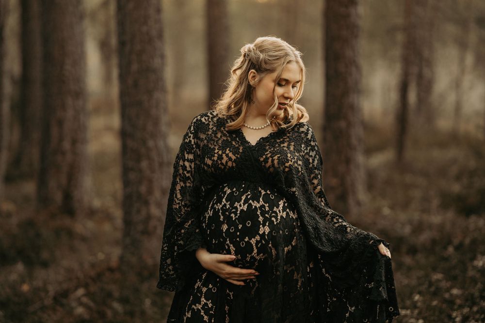 A pregnant figure in an elegant black lace dress stands gracefully in a moody autumn forest setting.