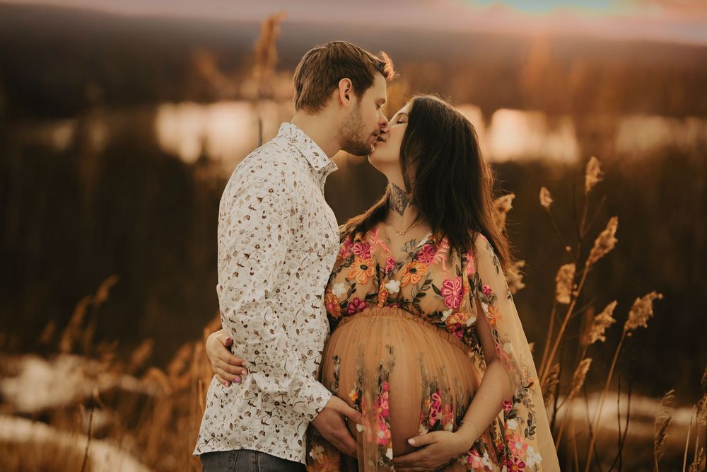 Expecting couple embraces in a field at sunset, with tall grass and a warm golden glow in the background.