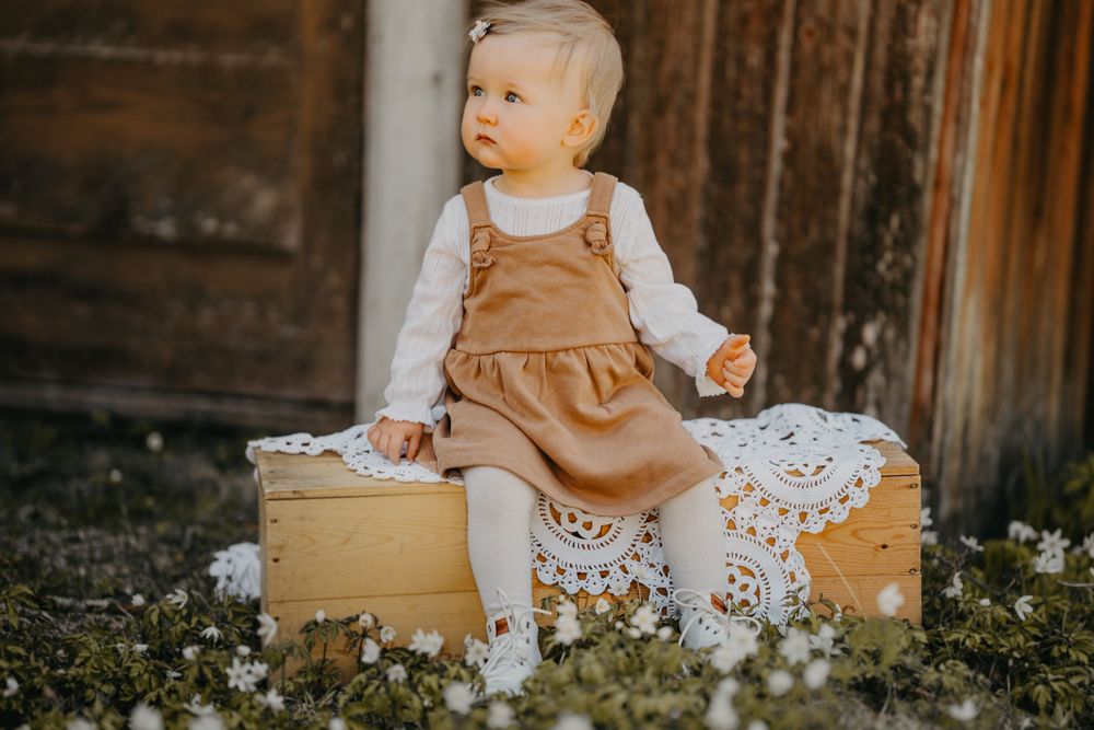 A small child wearing tan overalls and white tights sits on a rustic wooden box surrounded by delicate white flowers.
