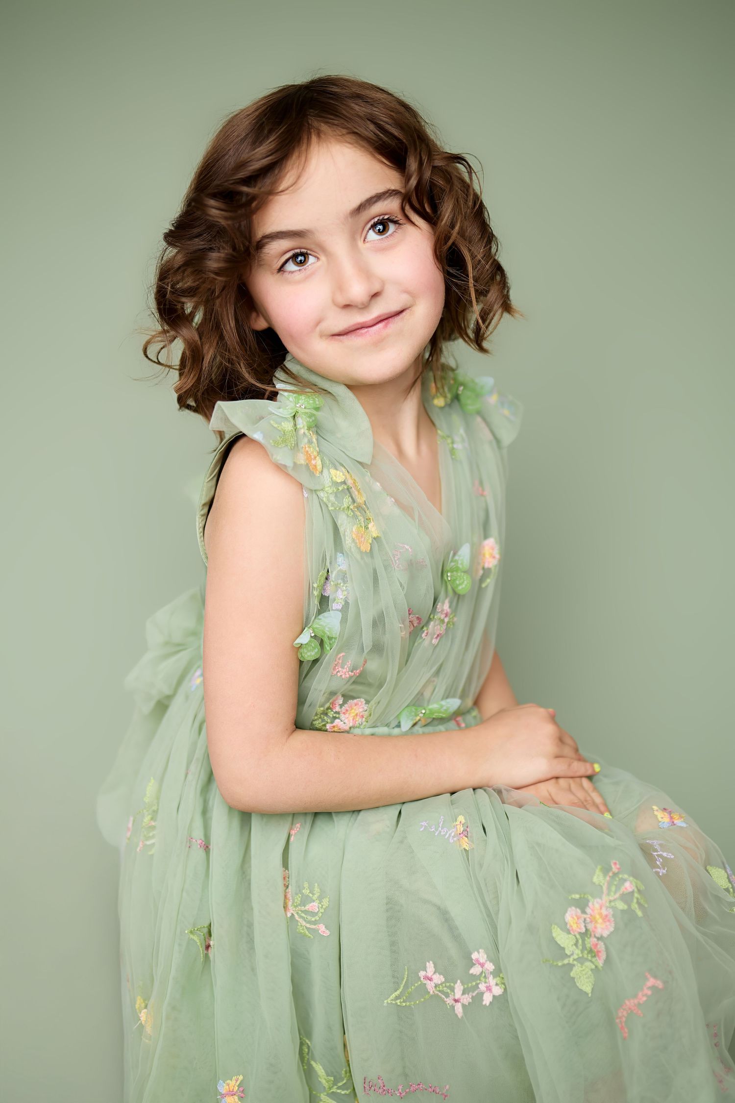 Young child in sage green floral dress twirls and poses playfully against mint green backdrop in portrait series.
