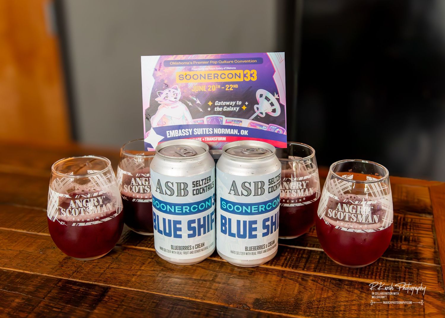 Cans of Blue Sky hard seltzer displayed with red-filled glasses and a promotional sign on a wooden table.