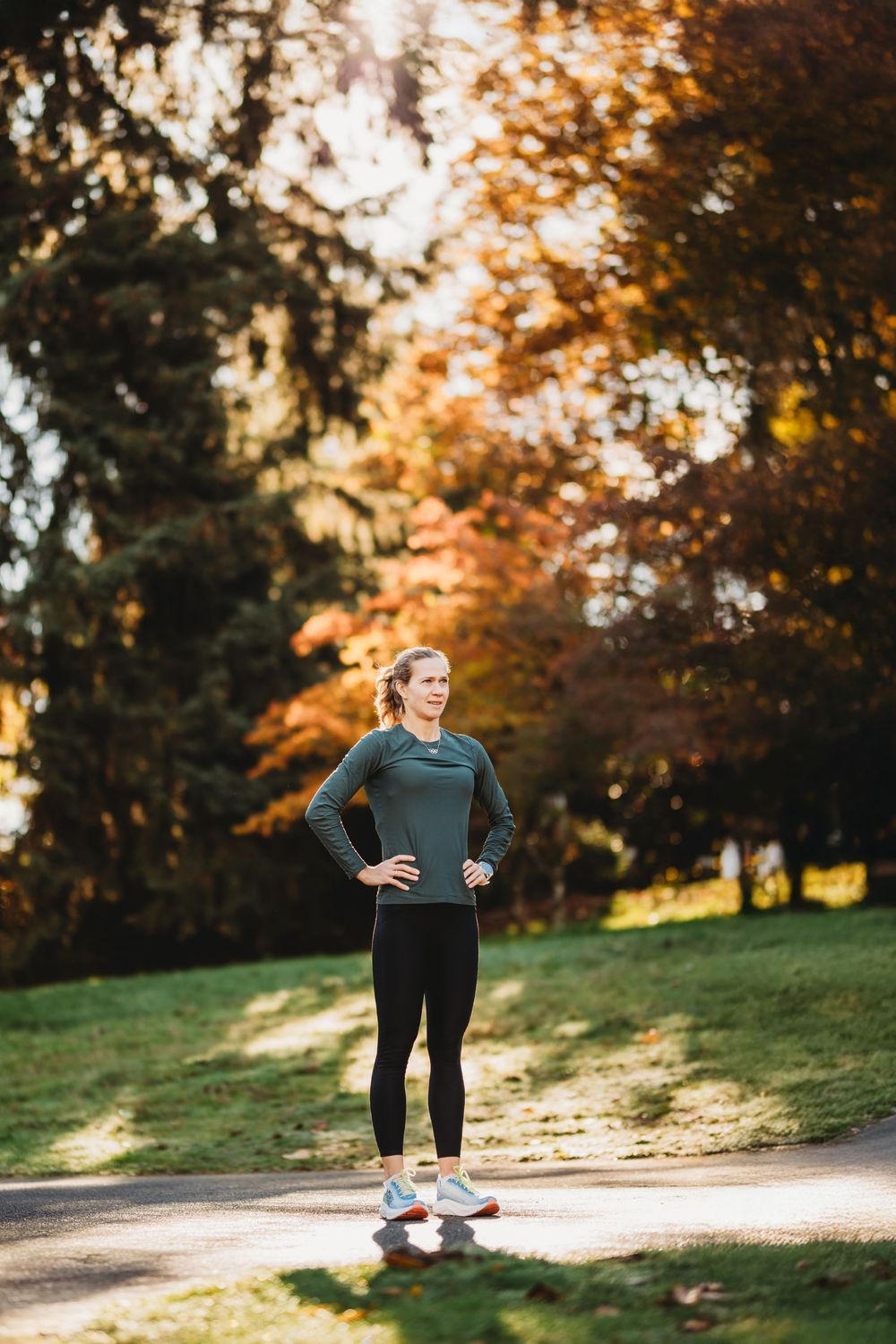 Athletic person in workout gear pauses on a sunlit path during autumn with colorful fall foliage in background.