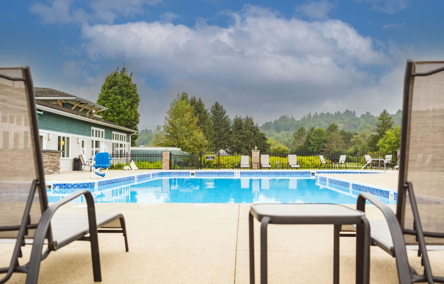 Outdoor swimming pool with lounge chairs at a resort surrounded by trees and blue sky on a sunny day.