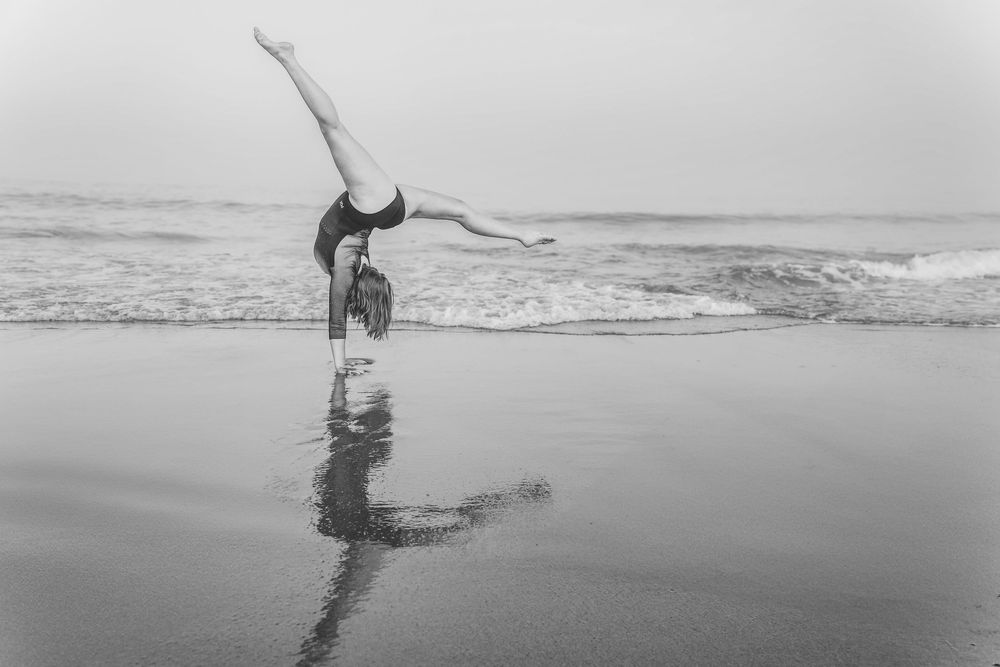 A high school senior and gymnast performs a graceful handstand on a reflective beach shoreline at dusk in black and white photography.