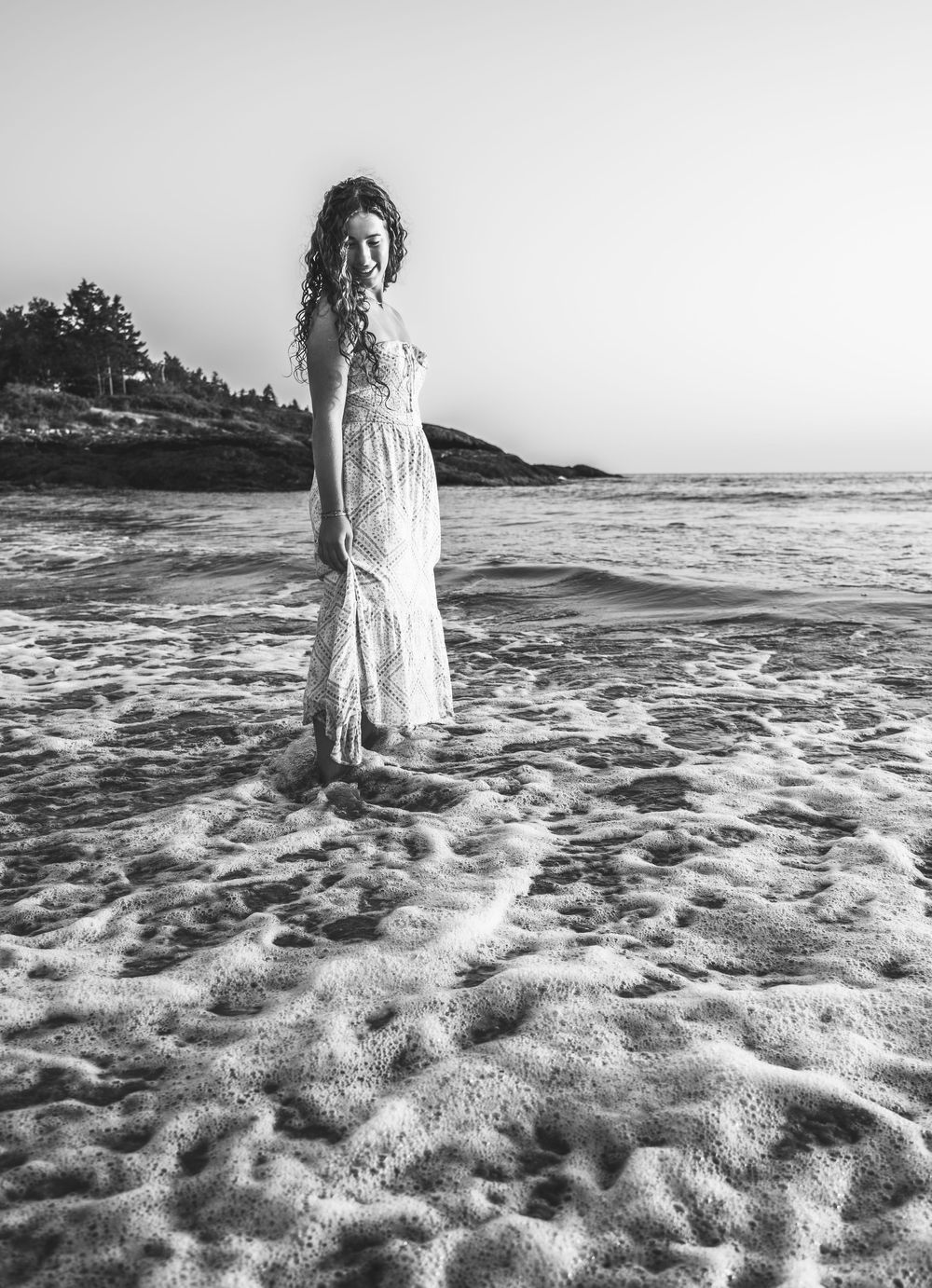 A rising senior in a flowy white dress stands at the edge of ocean waves on a sandy beach in black and white.