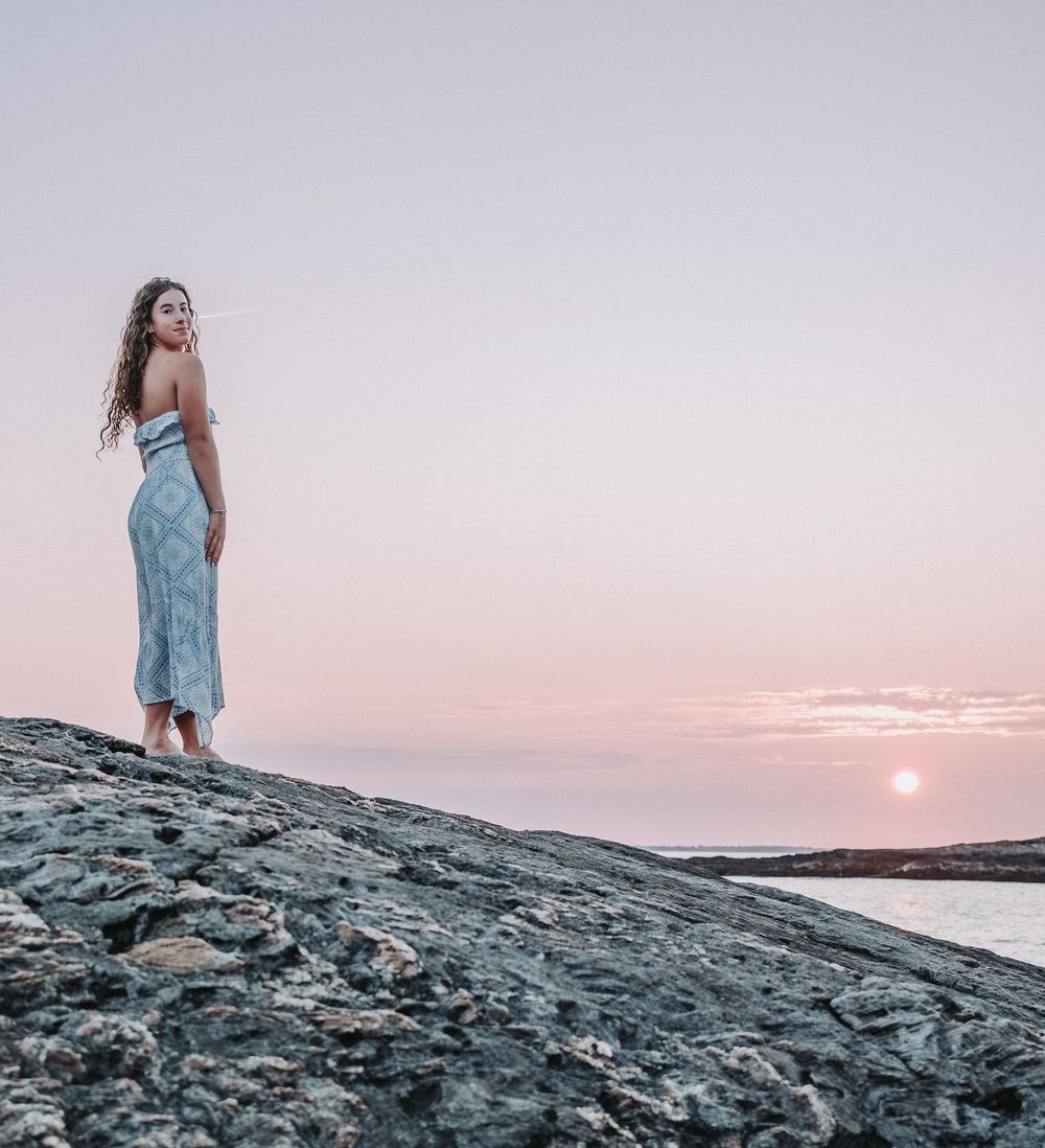 A high school senior stands on rocky coastal outcrop at sunset over ocean, wearing a white dress against pink and purple sky.