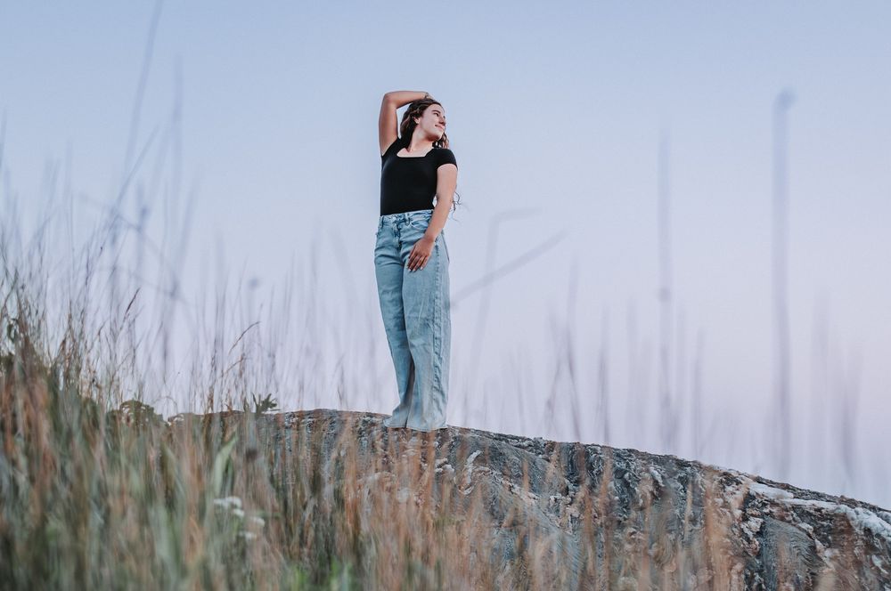 A rising HS senior, in blue jeans and a black shirt stands thoughtfully on a rocky hillside amidst tall grass on an overcast day.