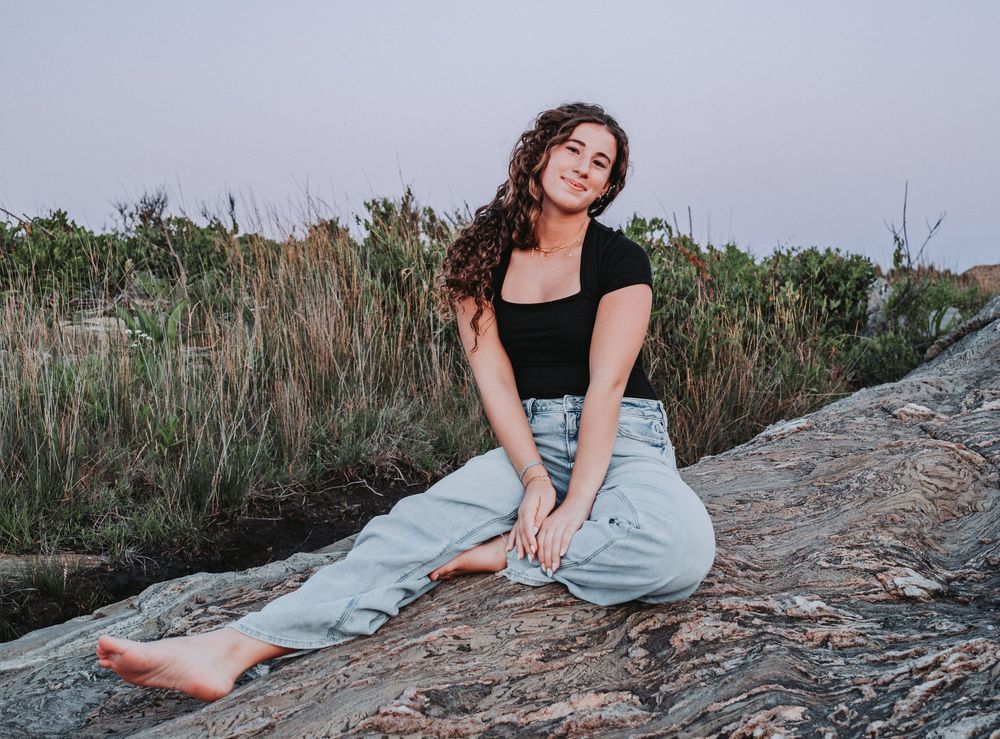A rising HS senior in a black top and light jeans sits  on a large rock at sunset with tall grass in the background at Hermit Island.