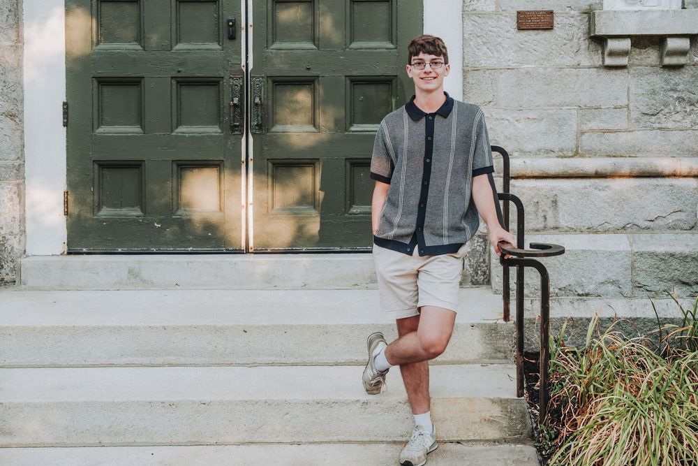 A person sits on a concrete step in front of a building entrance wearing casual clothing and sneakers.