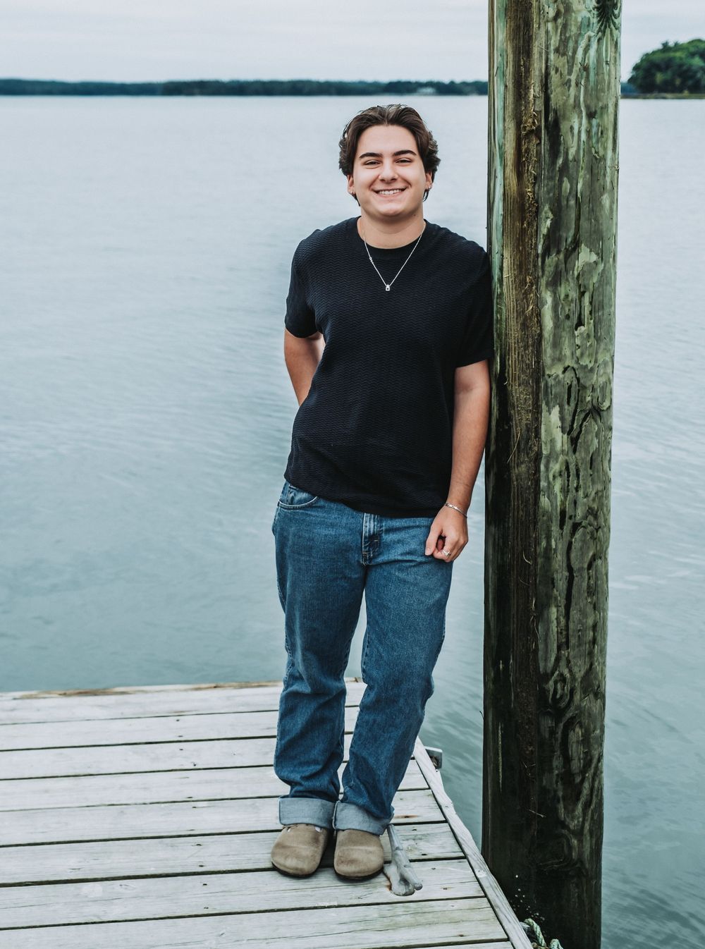 A senior from Freeport High School stands on a wooden dock by a calm water at Winslow Park wearing a black t-shirt and jeans.