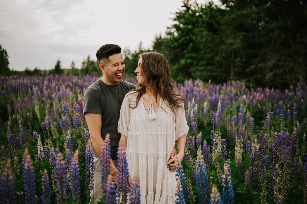 Fun and romantic portrait of couple looking at each other and laughing in lupine wildflower field Vancouver Island adventure Photographer