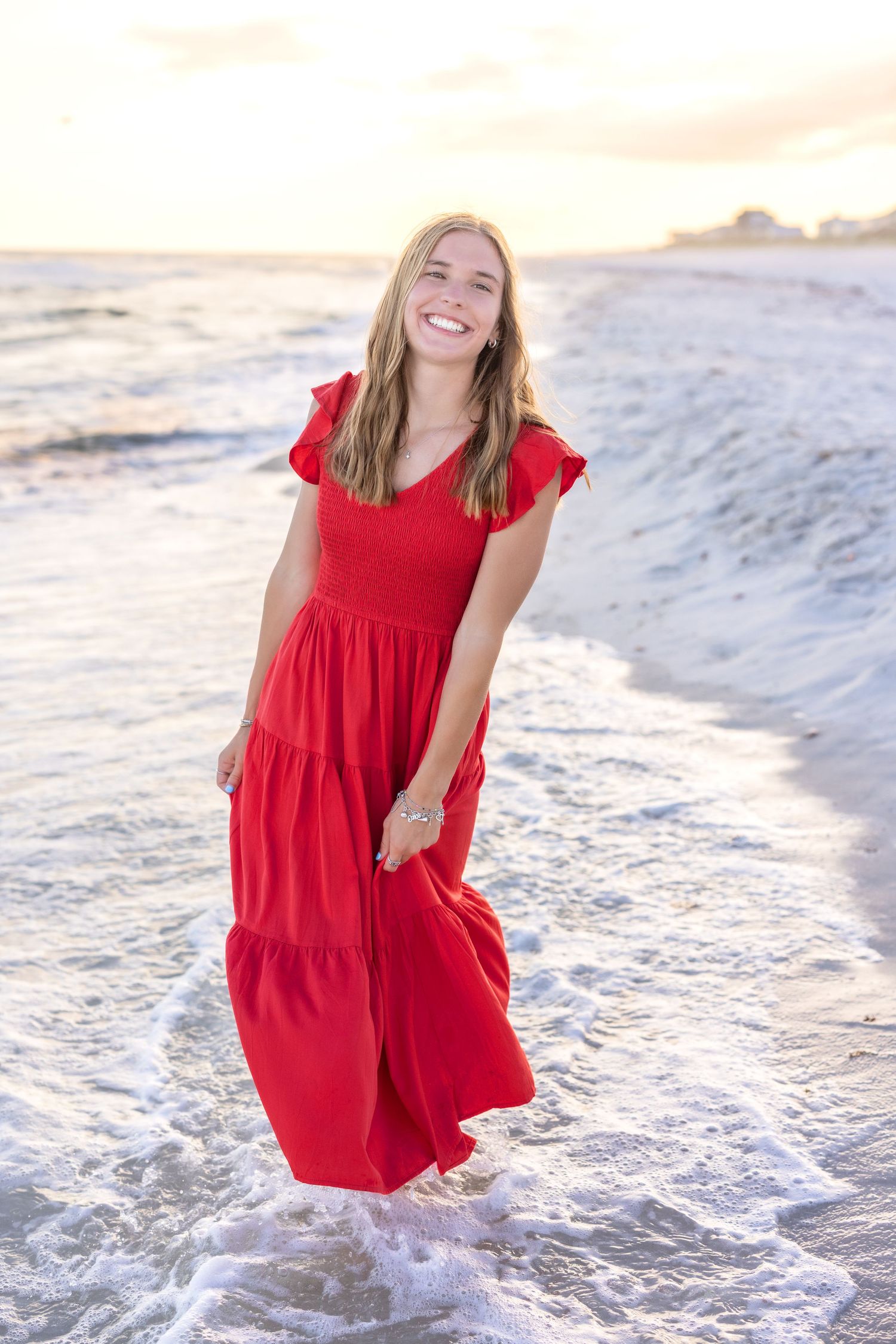 A person in a flowing red dress plays in ocean waves at sunset on a sandy beach.
