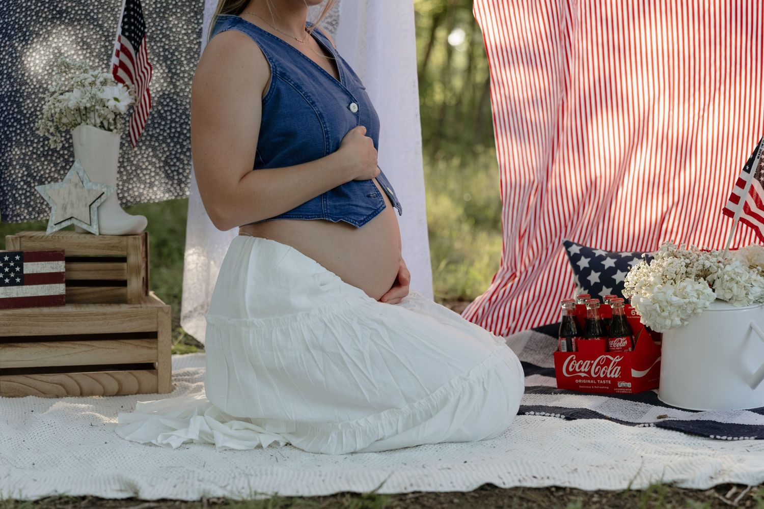 Outdoor picnic setting with white blanket, flowers in vase, and classic Coca-Cola bottle on grass in sunlight.