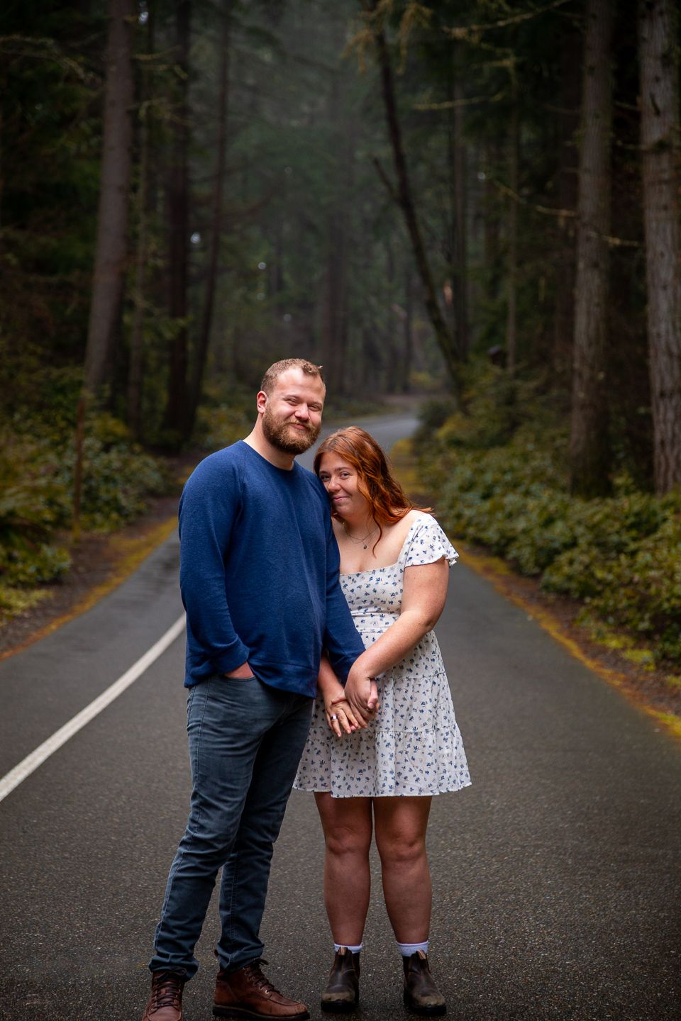 A couple embraces on a tree-lined asphalt road, wearing a navy blue shirt and white polka dot dress.