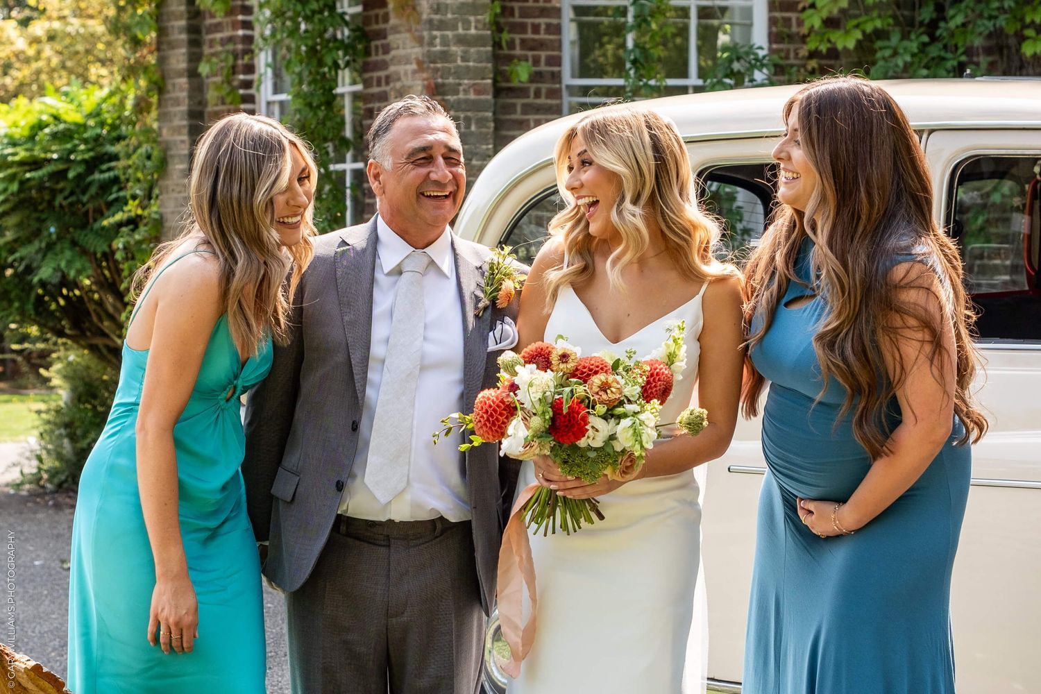 Wedding party laughing together outdoors in turquoise and blue bridesmaid dresses next to vintage white car.
