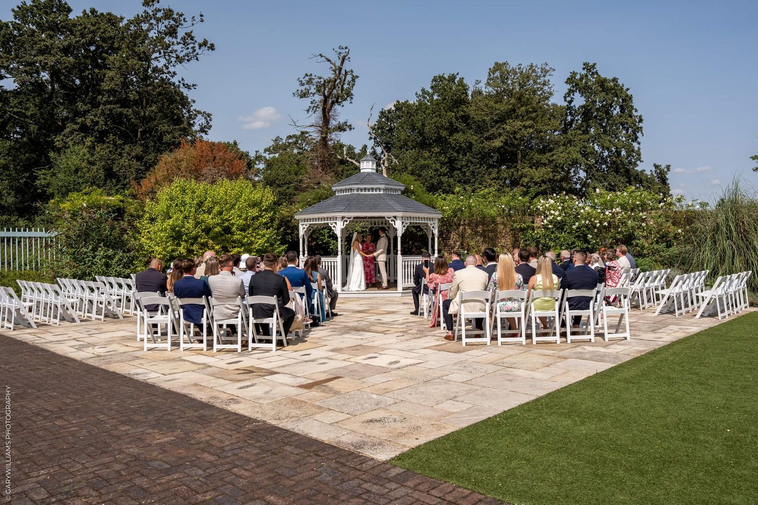 Outdoor wedding ceremony setup at a garden venue with white chairs and gazebo surrounded by lush greenery.