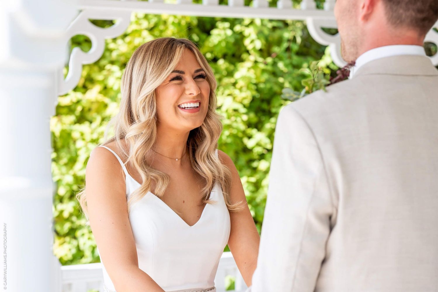 A bride in a white V-neck wedding dress smiles during an outdoor ceremony under natural greenery.