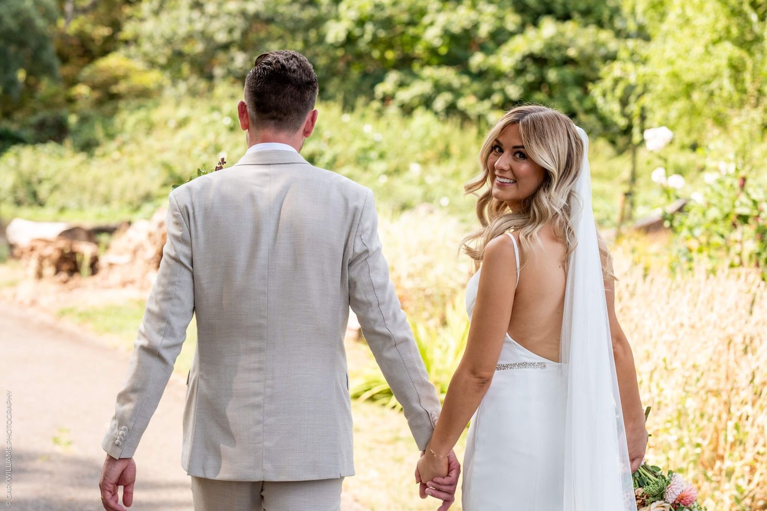 A bride and groom walk hand in hand down a path surrounded by golden sunlit foliage during an outdoor wedding.