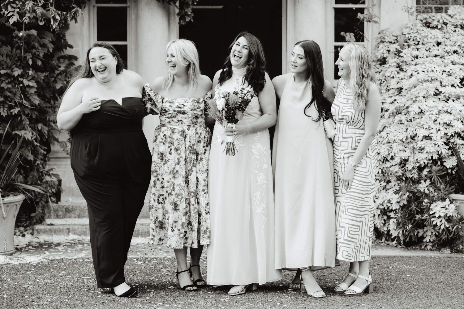 A group of women in formal dresses share a candid, joyful moment together on a lawn outside.