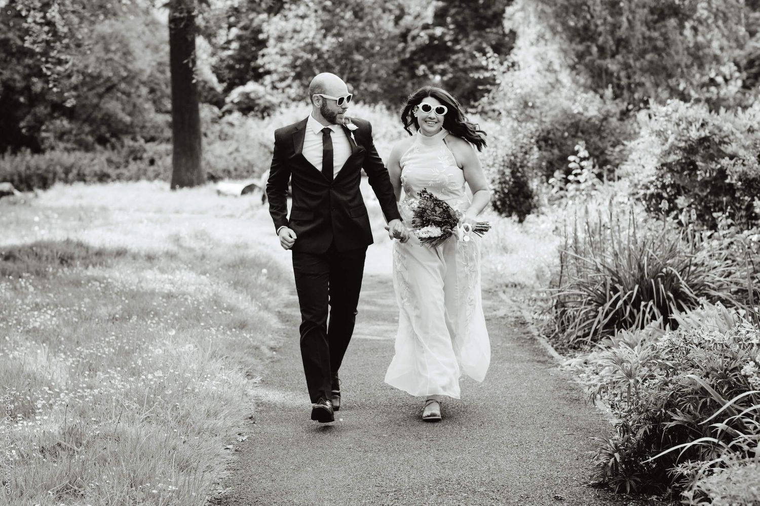 A couple walks together on a garden path surrounded by lush greenery in black and white photography.