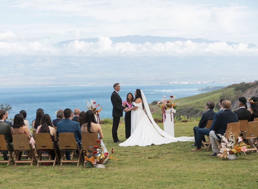 An outdoor wedding ceremony takes place on a scenic coastal hilltop with guests seated on wooden chairs overlooking the ocean.