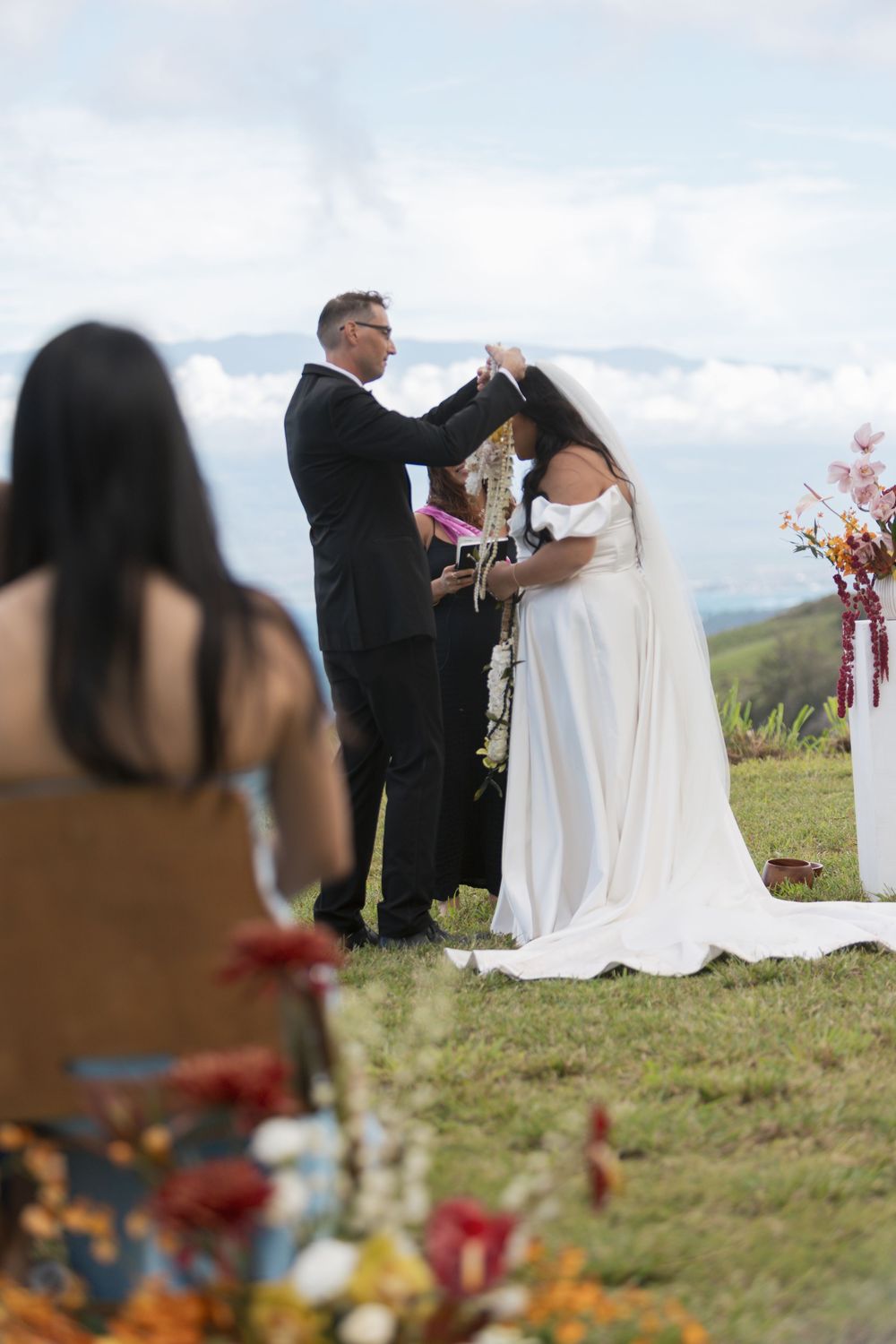 An outdoor wedding ceremony takes place on a hilltop with vibrant flowers in the foreground and misty mountains in the background.