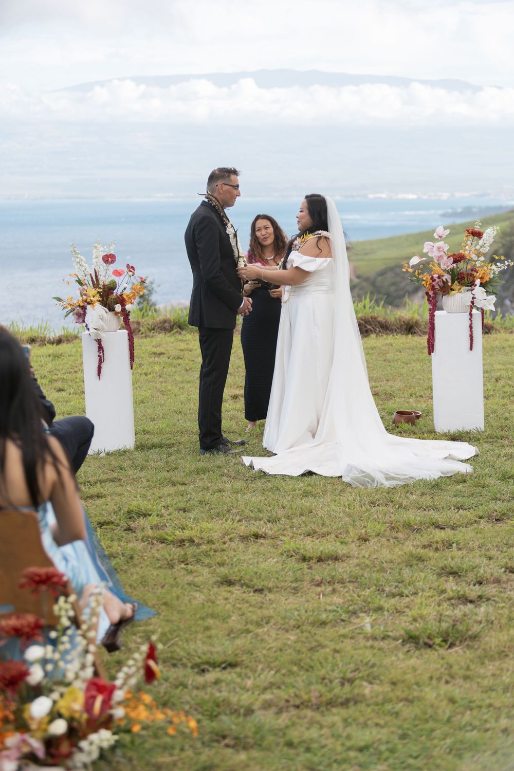 A scenic outdoor wedding ceremony on a hilltop with floral decorations on white pillars and an ocean view backdrop.
