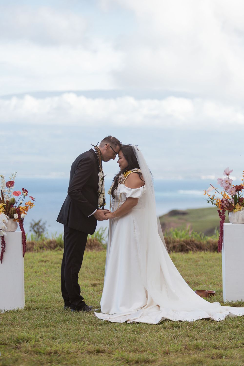 A romantic outdoor wedding ceremony with a couple exchanging vows surrounded by floral arrangements against a cloudy sky.
