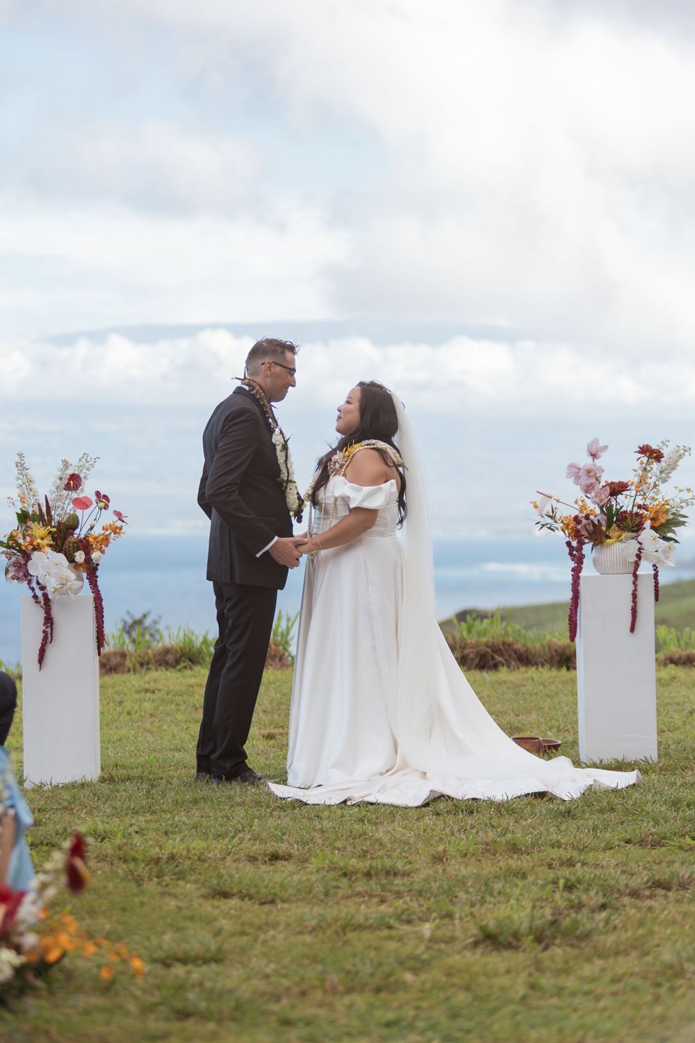 A couple shares an intimate moment during their outdoor wedding ceremony with floral arrangements on white pedestals.