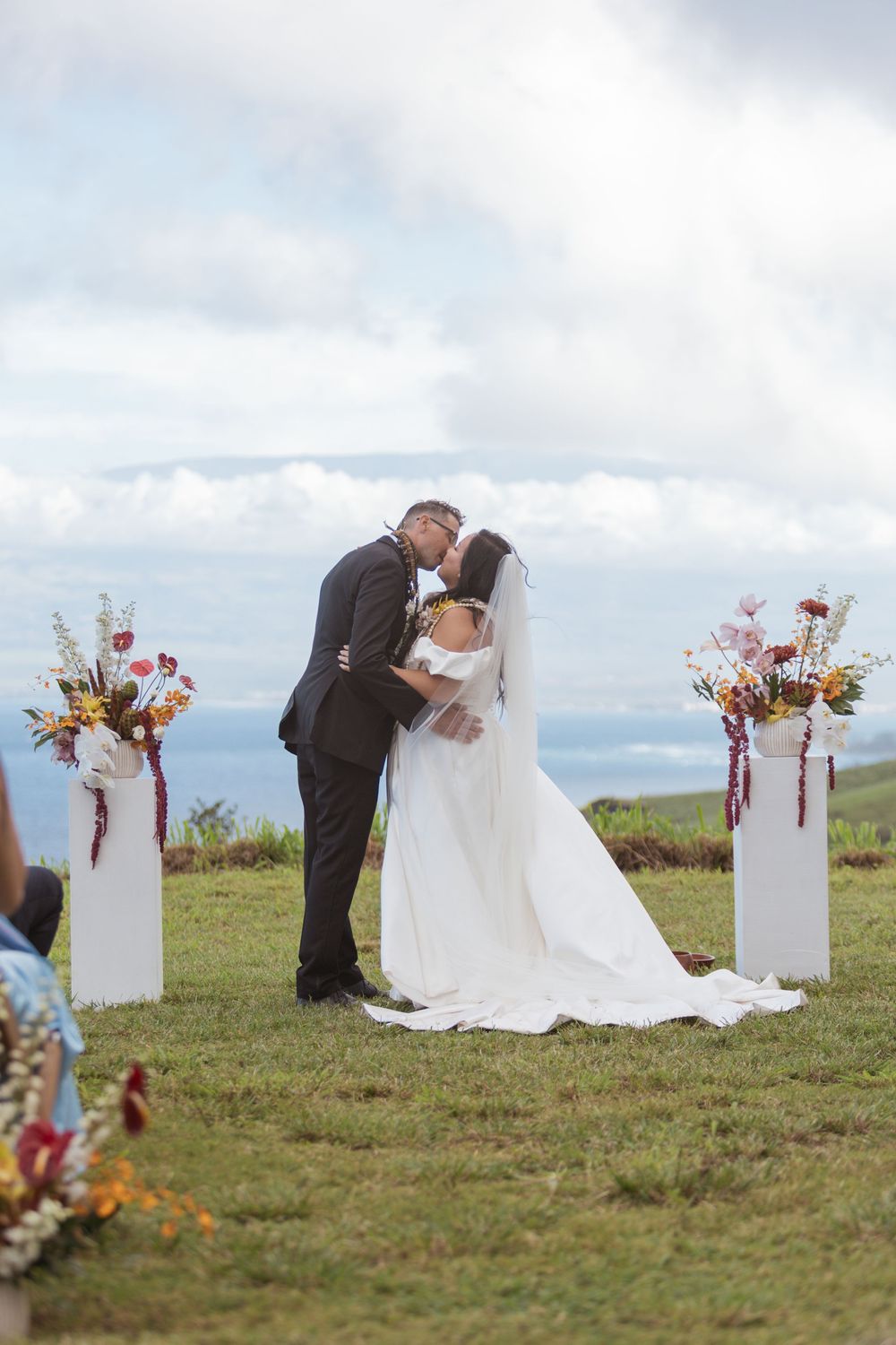 Romantic outdoor wedding ceremony on a grassy hilltop with floral pedestals against a cloudy sky backdrop.