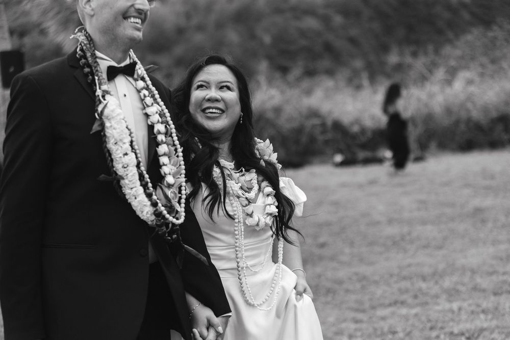 A happy couple wearing wedding attire and leis share a joyful moment outdoors in black and white photography.
