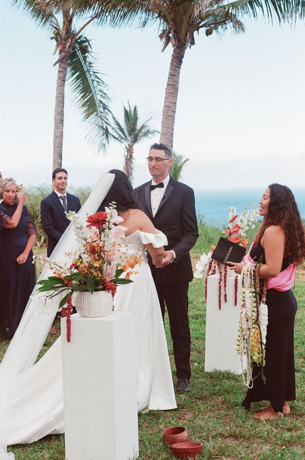A tropical wedding ceremony setup with palm trees and floral arrangements on white pedestals near the ocean.