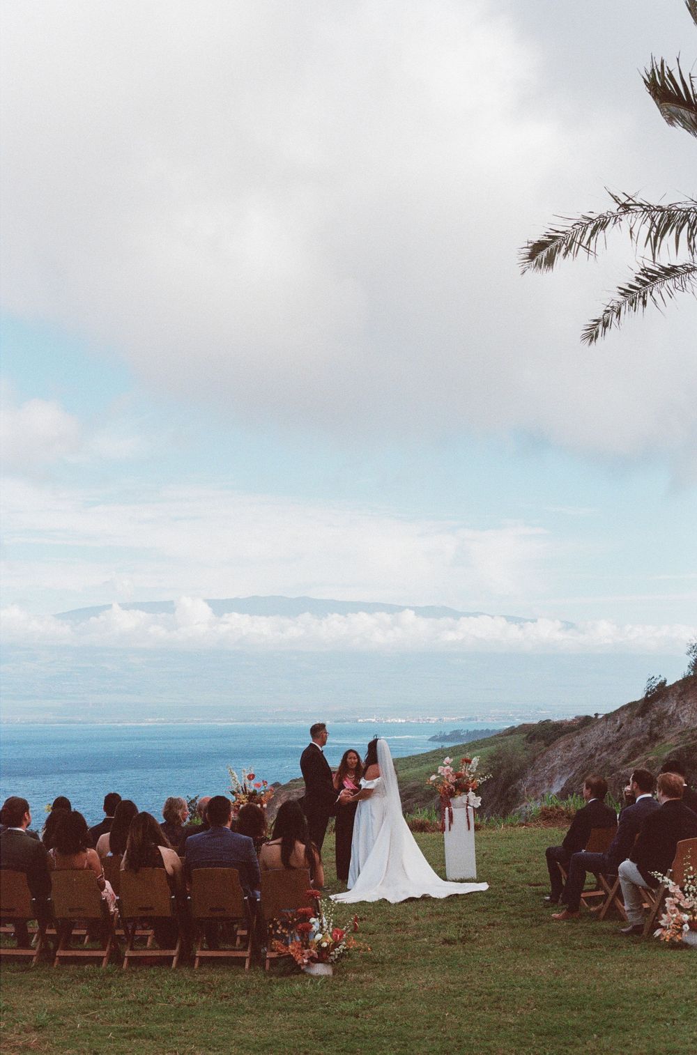 An outdoor wedding ceremony on a clifftop overlooking the ocean with wooden chairs and palm trees in the background.