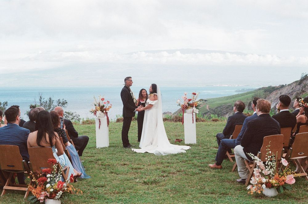 Panoramic coastal wedding ceremony overlooking the ocean with brown wooden chairs and floral arrangements.