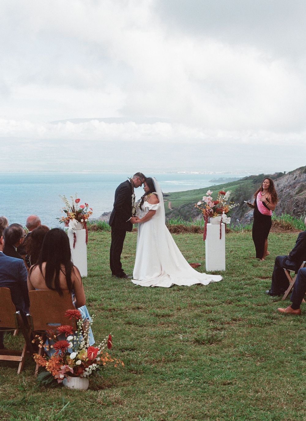 Outdoor cliffside wedding ceremony with floral arrangements and wooden chairs overlooking the ocean on a cloudy day.