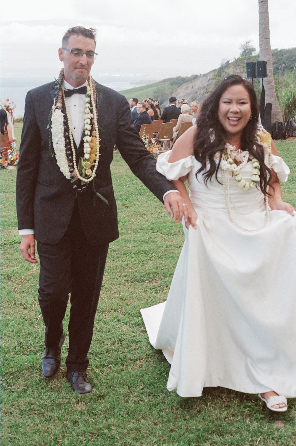 Wedding couple wearing traditional leis and formal attire celebrate outdoors on a grassy cliff overlooking the ocean.