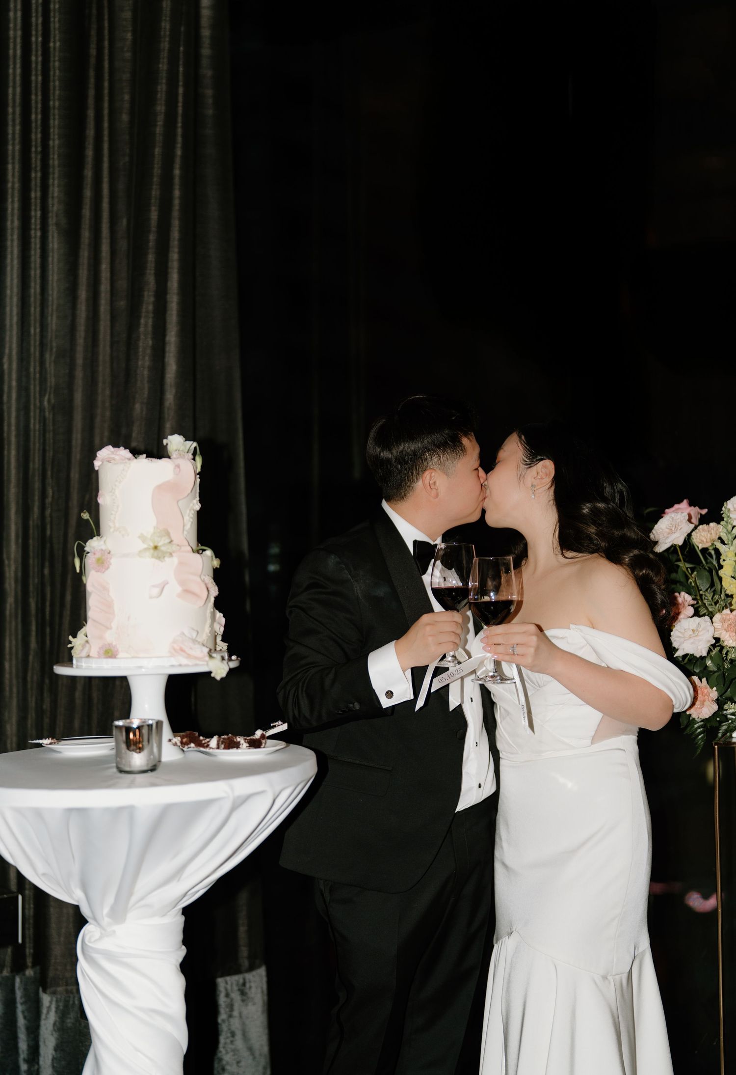 A couple shares an intimate moment while toasting next to an elegant three-tiered white wedding cake on a draped table.