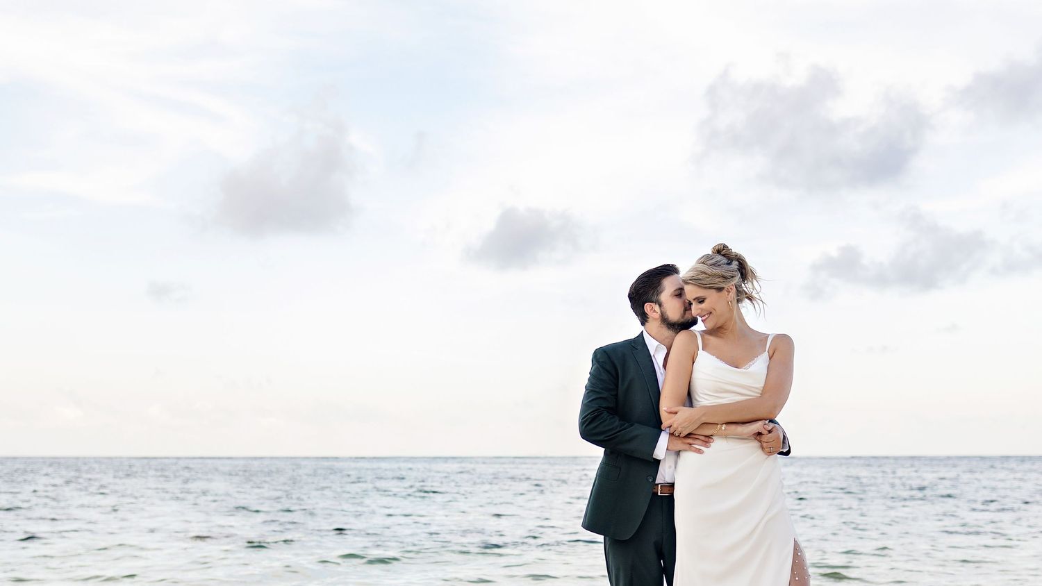 A couple in elegant wedding attire kisses and embraces on scenic beach steps during their destination ceremony.