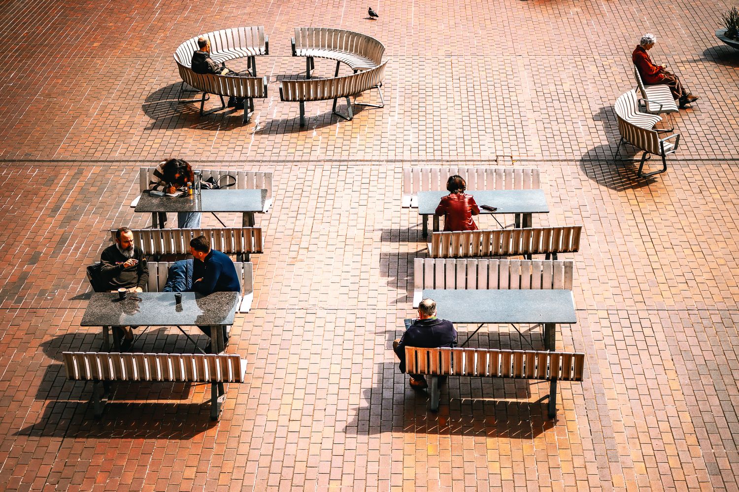 People seated at round tables in a sunlit brick-paved urban courtyard from above. The Barbican, City of London