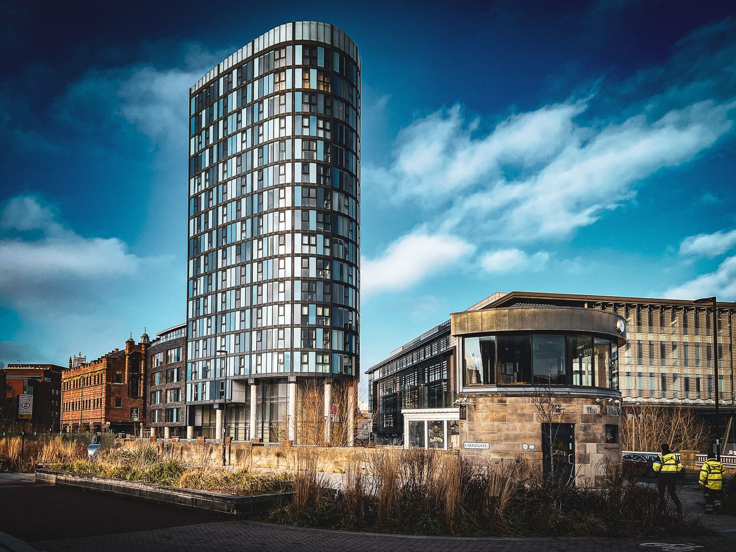 Crucible Stack, Sheffield. Modern cylindrical glass tower beside low-rise brick buildings and rough grassland.