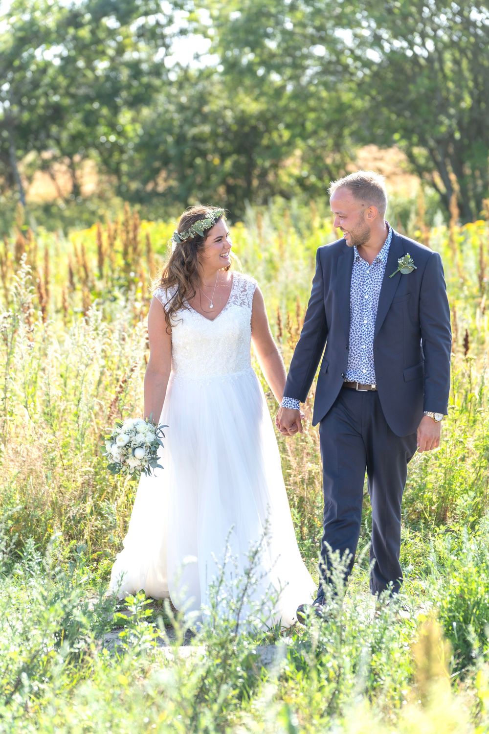 Couple in wedding attire holding hands while walking through a sunlit meadow with wildflowers and trees.