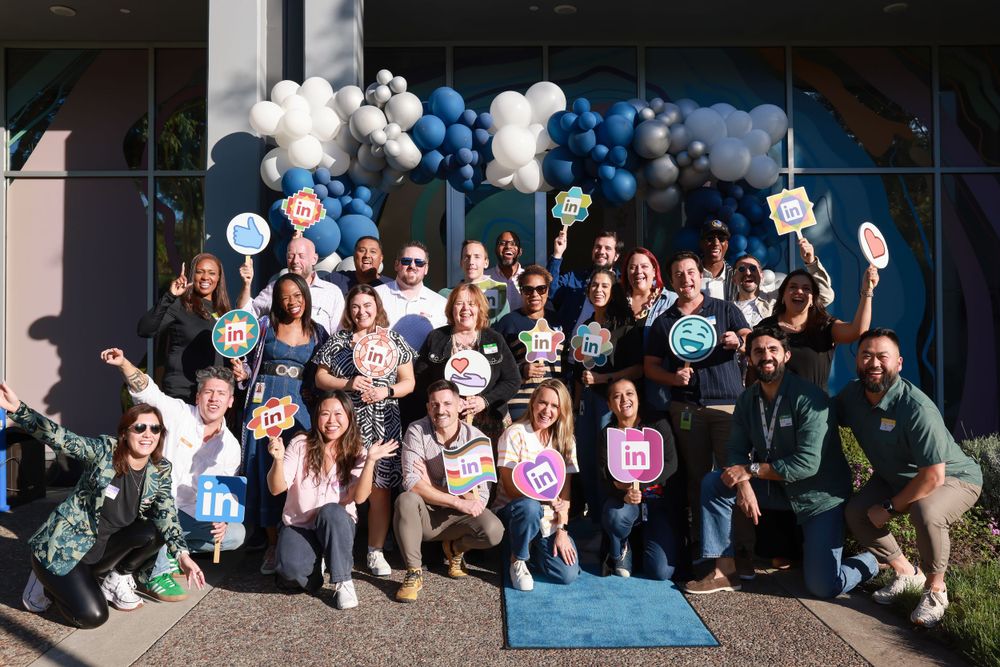 Large group of smiling employees holding social media logo signs in front of a balloon arch at LinkedIn event.